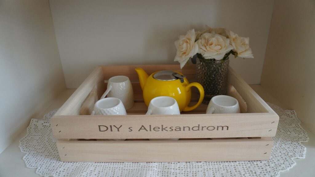Gajbica sa natpisom DIY s Aleksandrom, žutim čajnikom i šoljicama na belom kredencu.