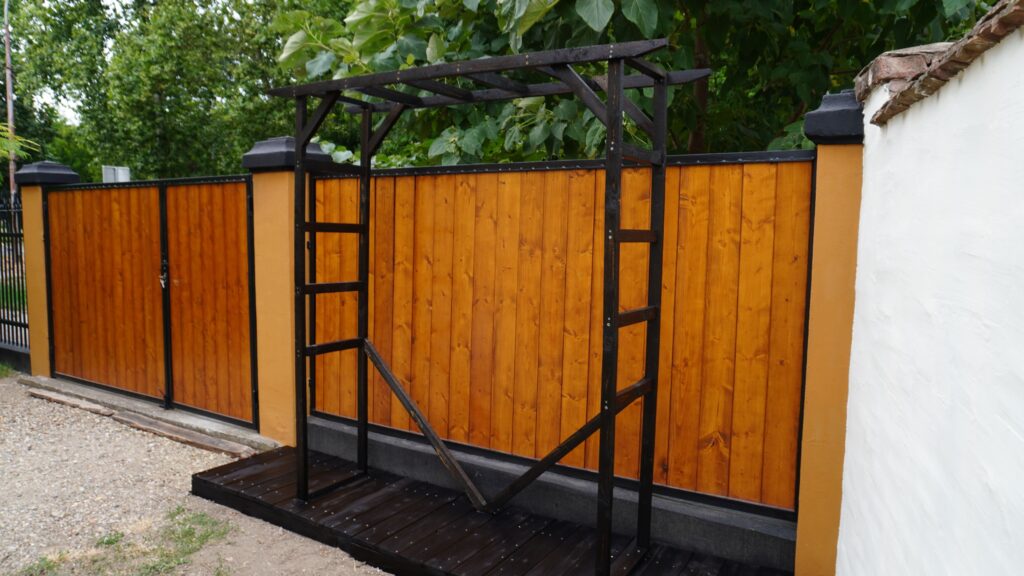 Tamna drvena mini pergola sa bočnim prečkama i donjim kosnicima uz ogradu