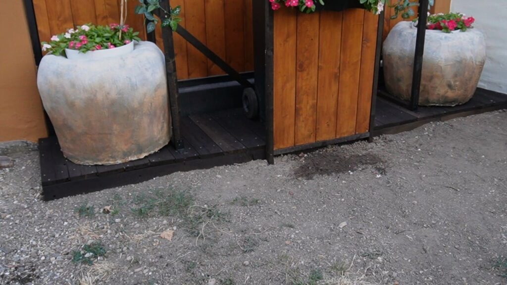 Manje saksije sa cvećem postavljene unutar dve dekorativne žardinjere na drvenoj platformi